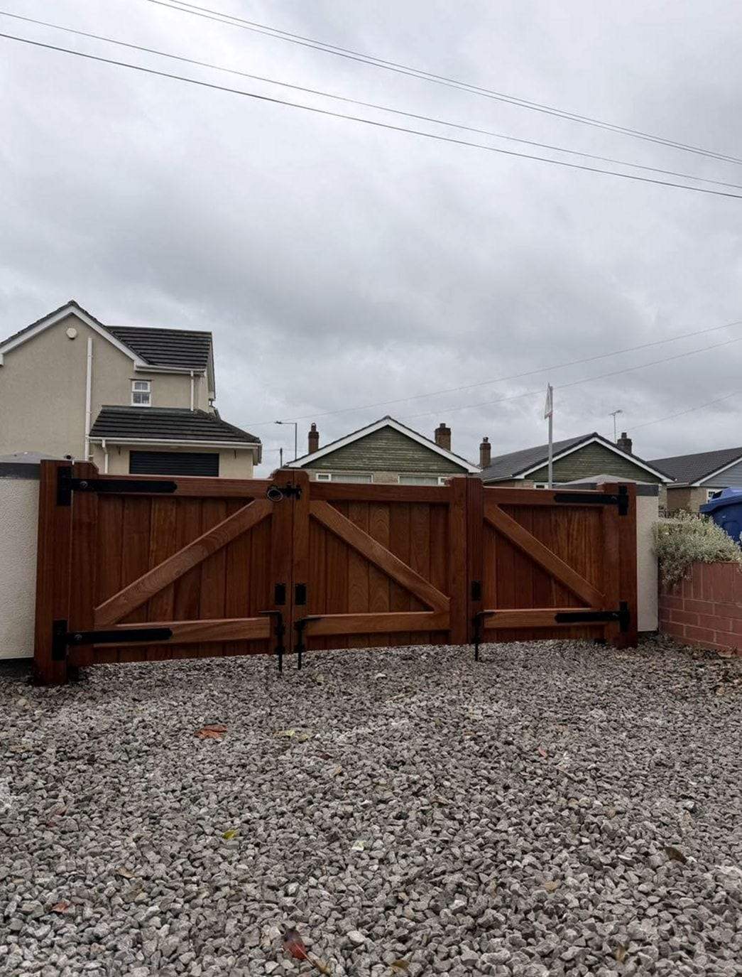 Pair of stained hardwood driveway gates with diagonal Z-braces, black strap hinges and drop bolts, closed across a gravel drive