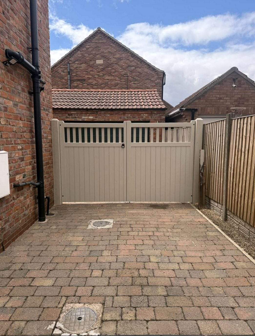 Pair of taupe painted gates with a slatted band along the top, set between a brick wall and a wooden fence at the end of a block-paved side drive