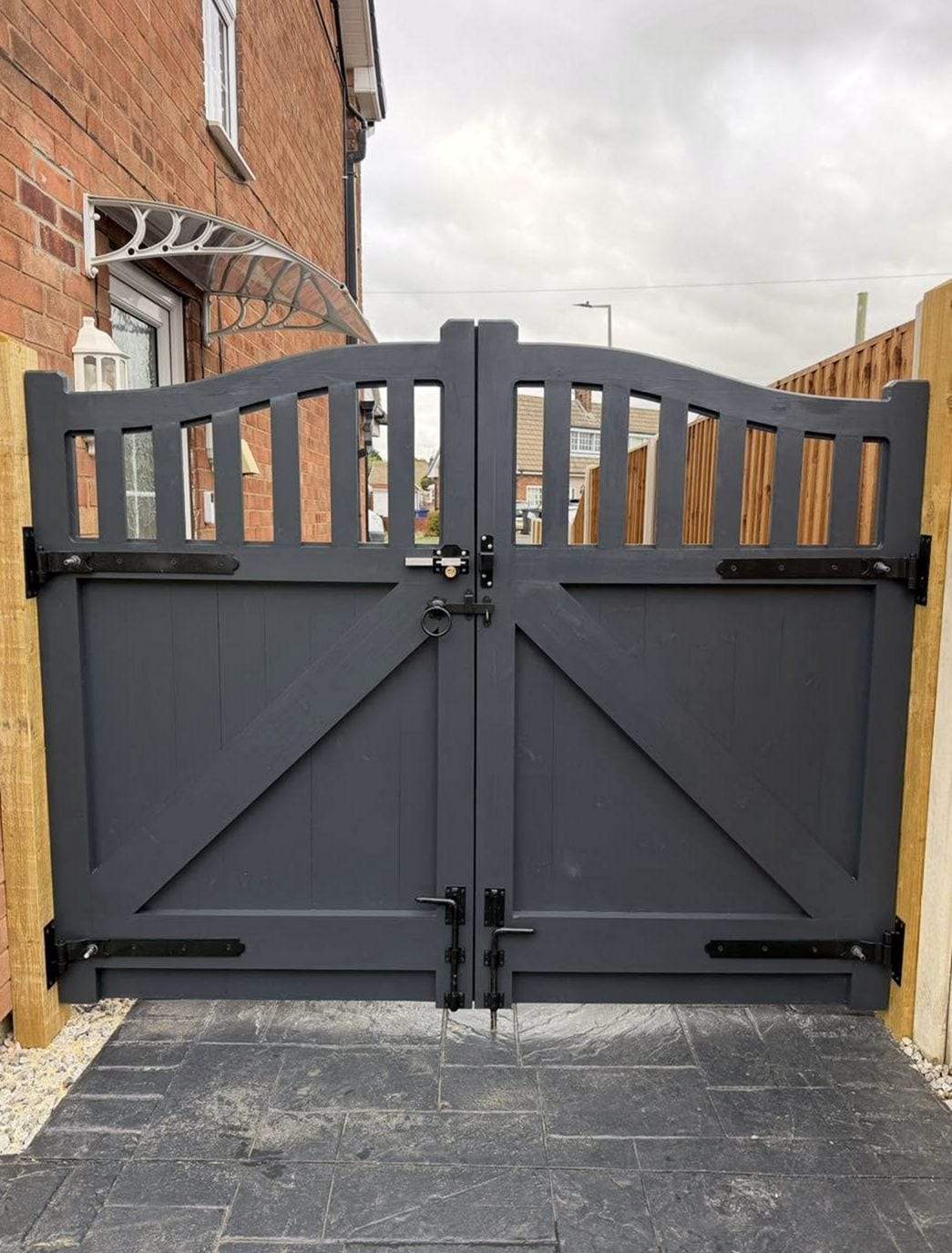 Pair of slate gray painted timber gates with an arched row of spindles on top, diagonal braces, black strap hinges, a latch and drop bolts, across a paved side entrance beside a brick house