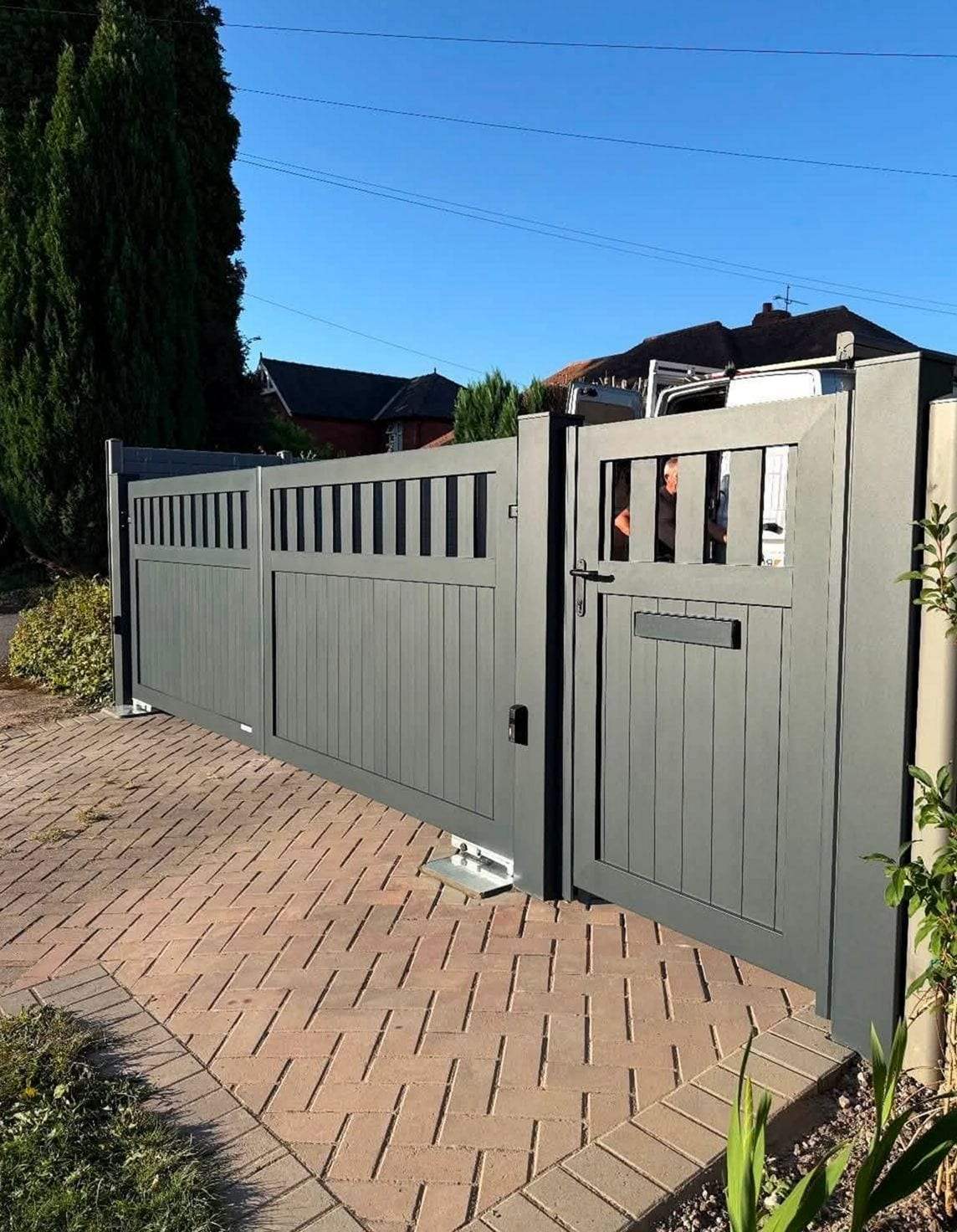 Anthracite gray sliding driveway gate with a slatted top band and a matching walk-through gate beside it, on a block-paved drive
