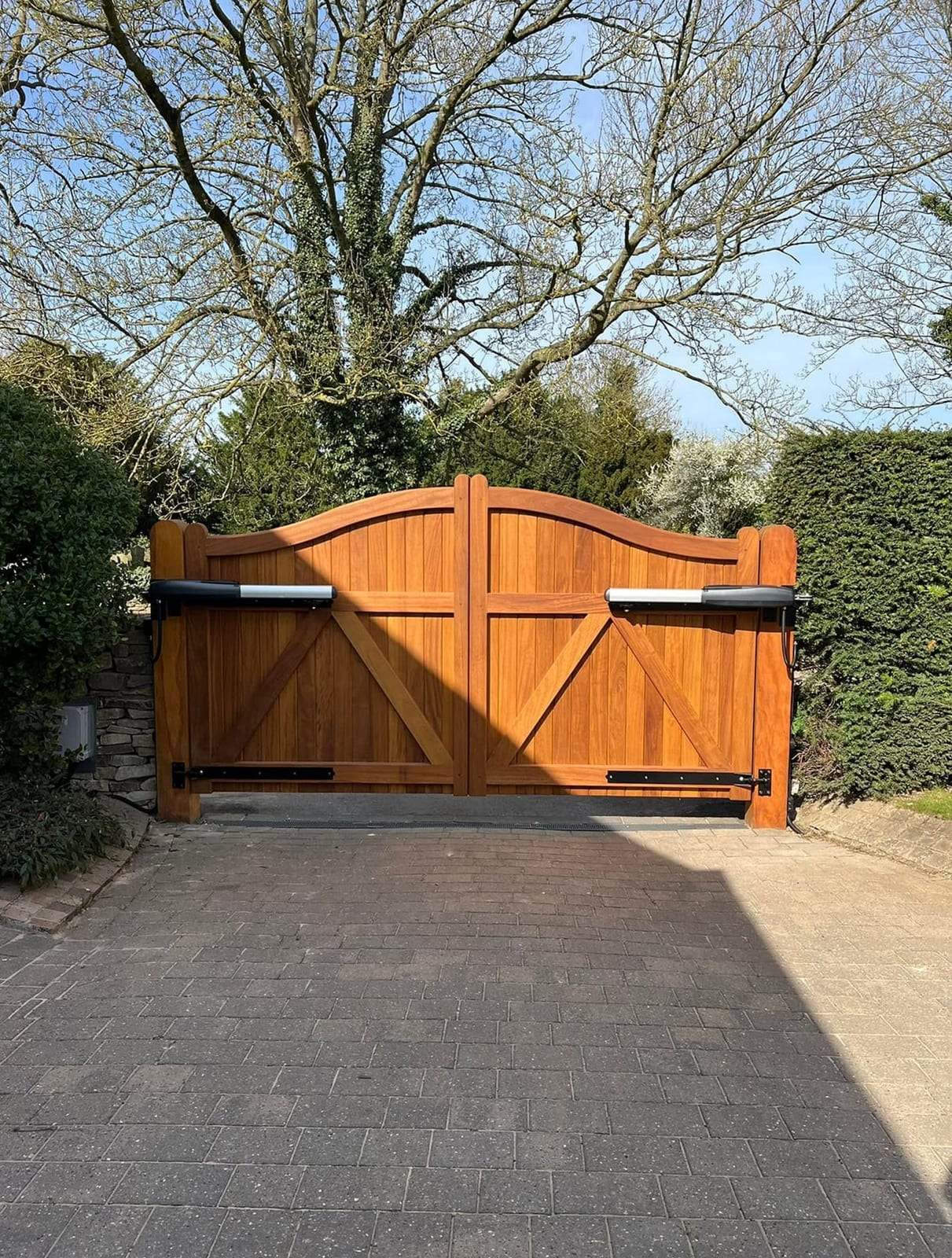 Arched hardwood double driveway gates with two ram-style automatic openers mounted along the top rail, closed across a paved drive under a large tree