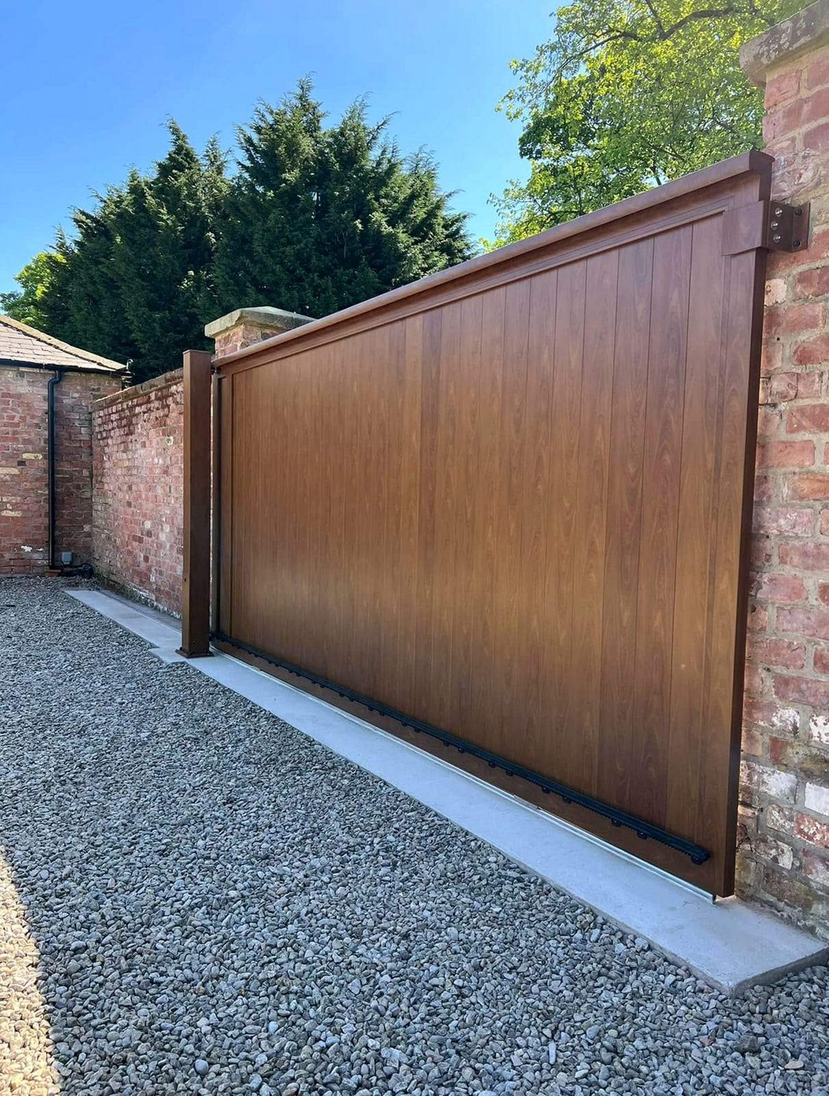 Tall wood-grain sliding gate running on a ground track along a concrete strip, between a weathered brick pier and a brick wall, beside a gravel drive