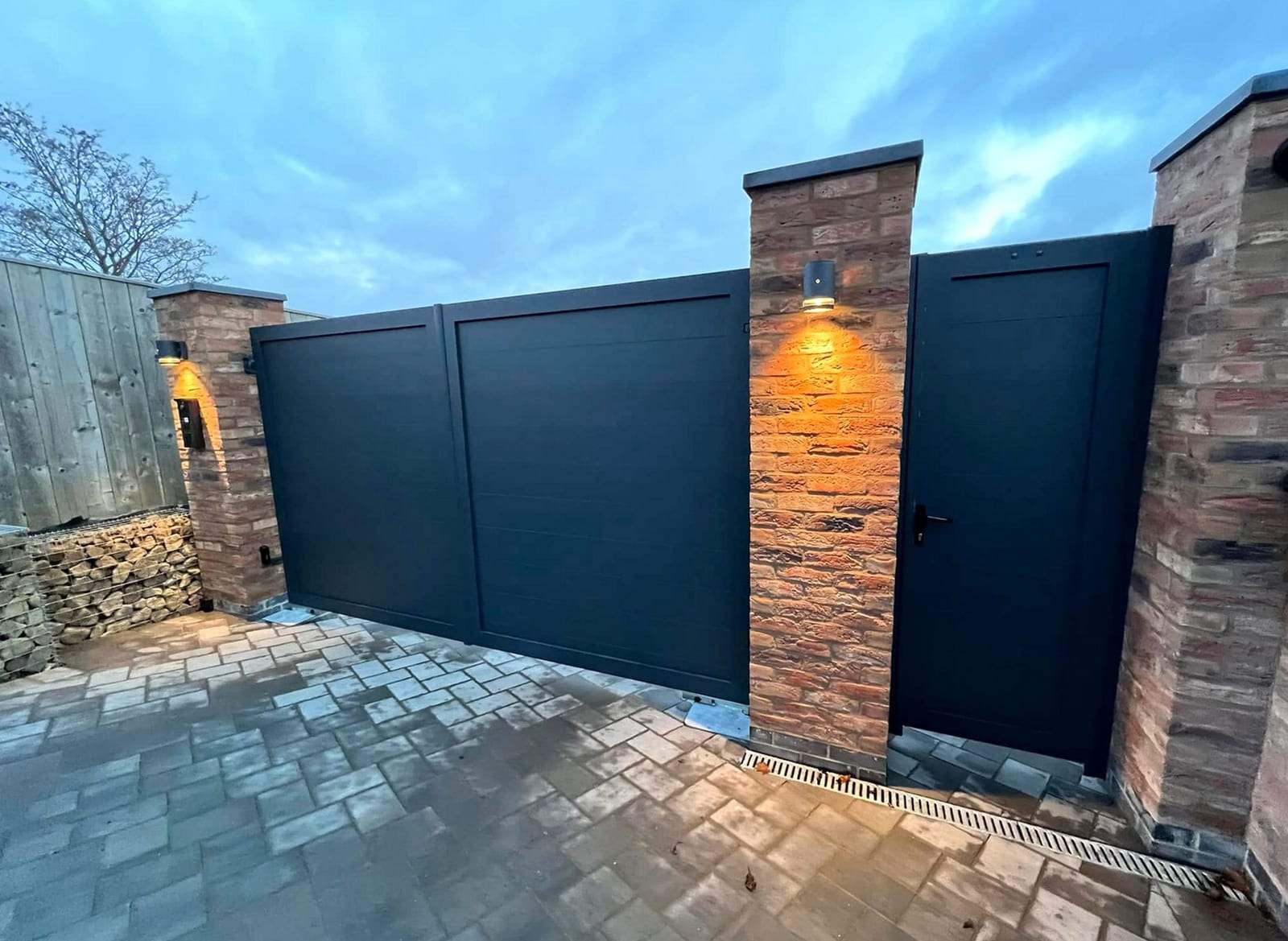Dark charcoal flat-panel double gates and a matching pedestrian door between brick piers, with wall lights glowing at dusk above a stone-paved entrance