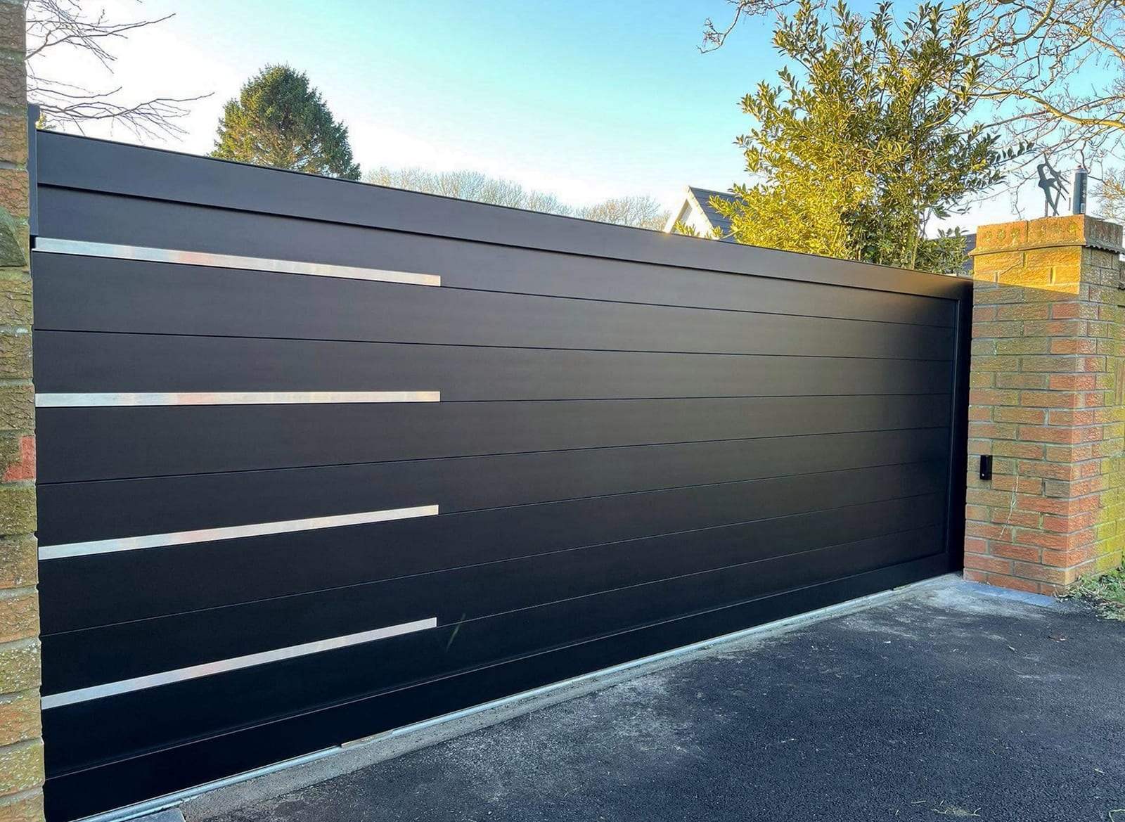Black sliding driveway gate built from horizontal panels with four brushed stainless accent strips, closed beside a brick pillar on an asphalt drive