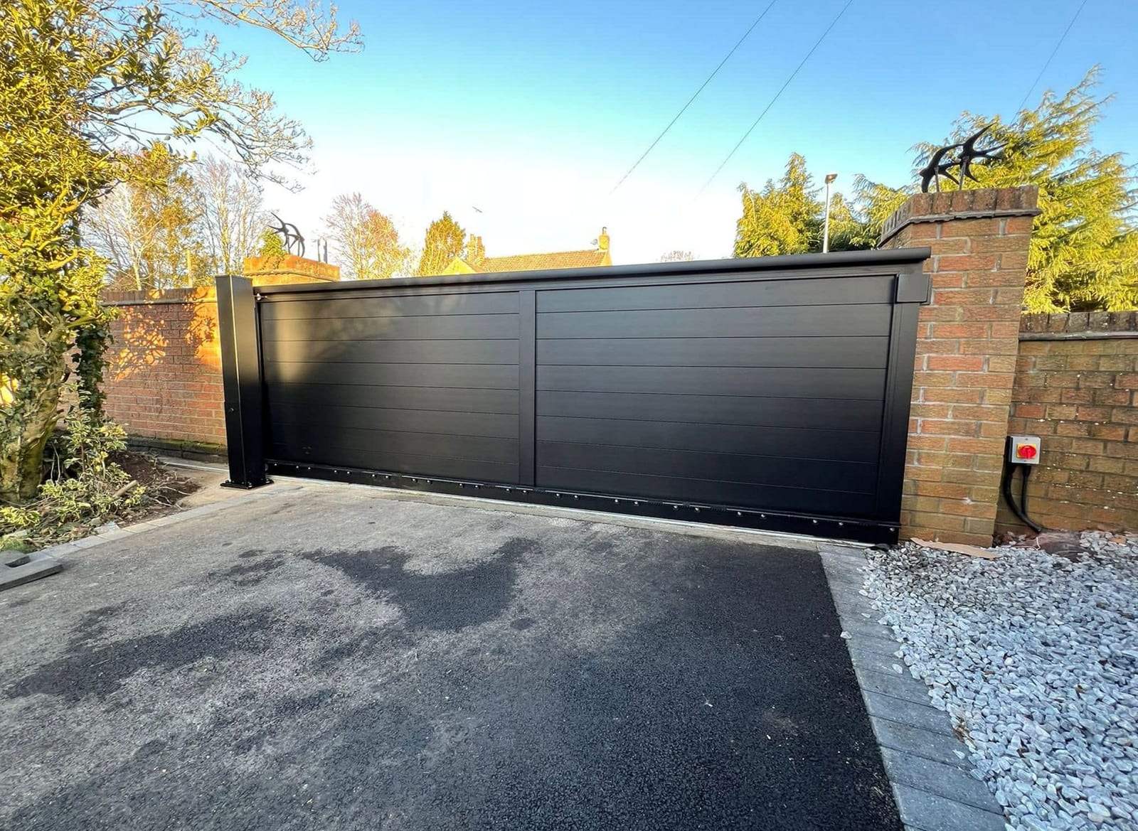 Black horizontal-panel sliding gate closed across an asphalt driveway, with a rotary power isolator switch on the brick wall beside it and its cables running down into the ground